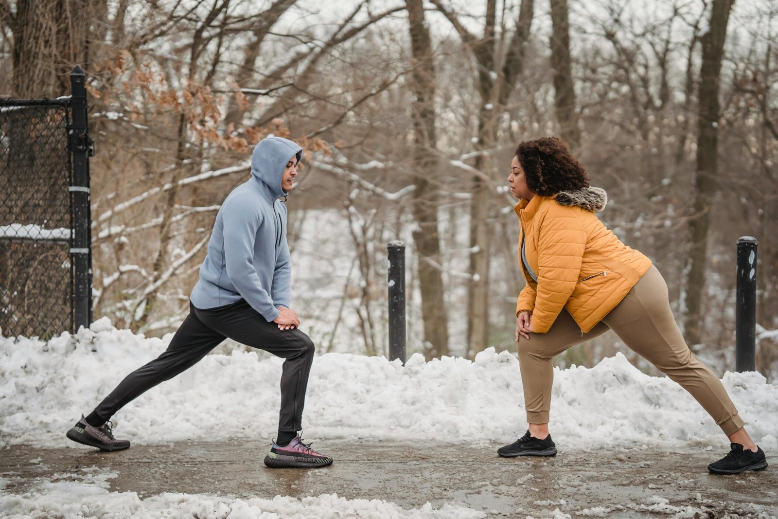 A couple performing warm-up stretches in a snowy outdoor park setting during winter.